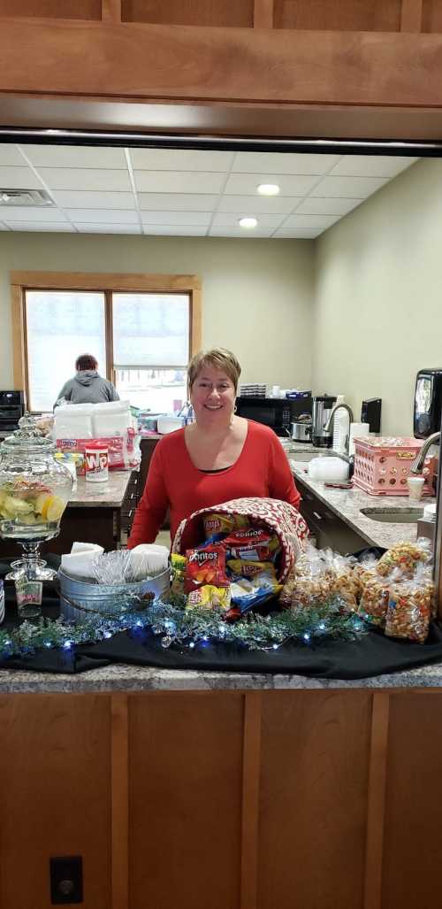 A woman stands behind a counter filled with festive snacks and treats, smiling in a cozy, decorated space.