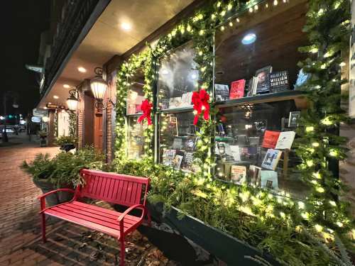A cozy bookstore window decorated with lights and greenery, featuring books and red bows, beside a red bench.