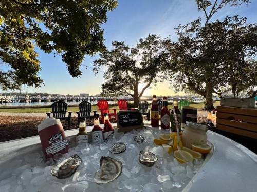 A seafood display with oysters, sauces, and lemons on ice, set against a scenic waterfront view at sunset.