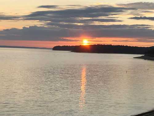 A serene sunset over calm waters, with vibrant colors reflecting on the surface and a silhouette of trees in the distance.