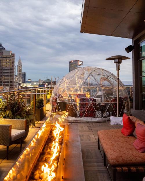 A rooftop terrace with a fire feature, cozy seating, and a transparent dome overlooking a city skyline under cloudy skies.
