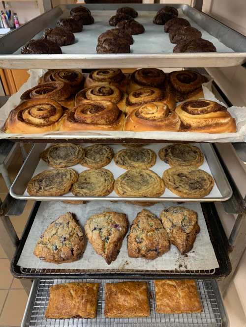 A bakery display with three trays of assorted baked goods: chocolate cookies, cinnamon rolls, and scones.