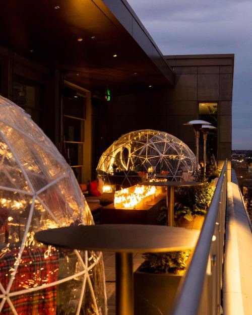 Two illuminated igloo-like structures on a rooftop terrace, surrounded by plants and outdoor heaters, at dusk.
