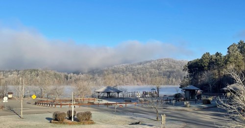 A serene winter landscape featuring a foggy lake, frosted trees, and a park with a gazebo under a clear blue sky.