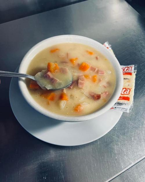 A bowl of creamy soup with diced ham and carrots, accompanied by a packet of crackers on a silver surface.
