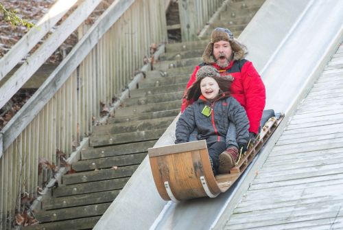 A man and a child joyfully ride a wooden sled down a snowy slope, both wearing winter clothing and hats.
