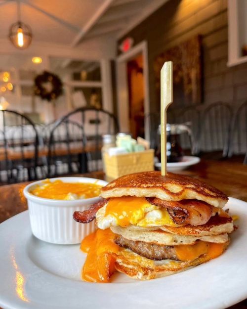 A stacked breakfast sandwich with eggs, bacon, and cheese, served with a side of creamy grits in a cozy café setting.