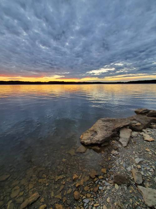 A serene lakeside view at sunset, with colorful clouds reflecting on the water and rocky shoreline in the foreground.