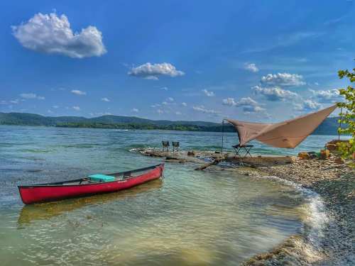 A red canoe rests on a sandy shore by a calm lake, with a tent and chairs set up under a blue sky with fluffy clouds.