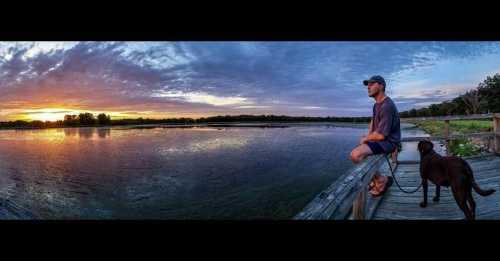 A person sits on a dock with a dog, watching a colorful sunset over a calm lake.