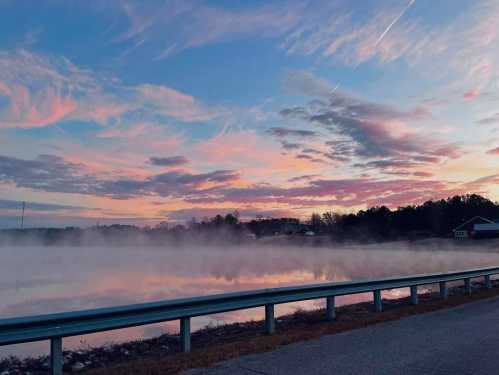 A serene landscape at dawn with mist over a calm lake, colorful clouds reflecting in the water, and a road in the foreground.