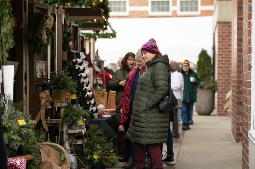 A woman in a green coat browses holiday decorations at an outdoor market, surrounded by festive stalls and shoppers.