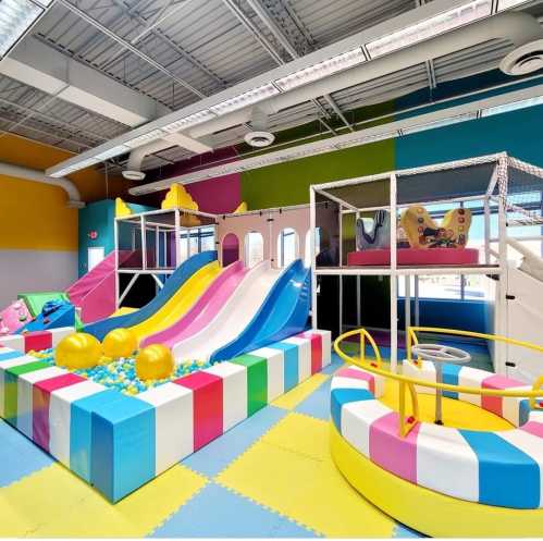 Bright indoor playground featuring colorful slides, ball pit, and a spinning ride, with soft flooring and vibrant walls.