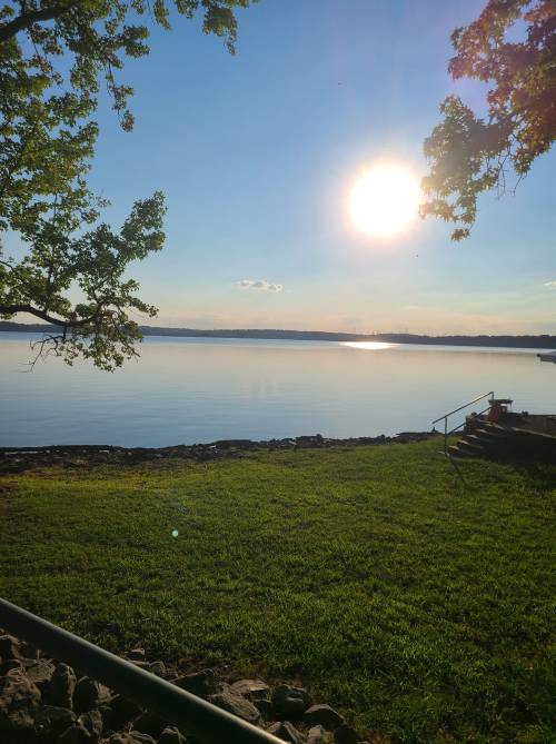 A serene lake view at sunset, with calm waters reflecting the sun and green grass in the foreground.