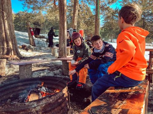 Three children sit by a fire pit in a snowy outdoor setting, enjoying the warmth and chatting together.