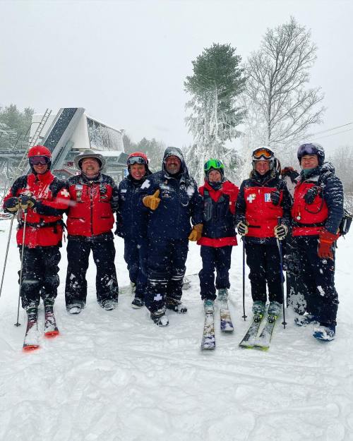 A group of seven skiers in red jackets stands together in a snowy landscape, smiling amidst falling snow.