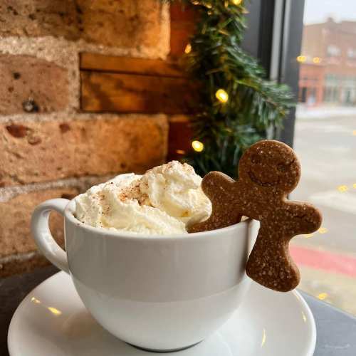 A white mug filled with whipped cream and topped with a gingerbread cookie, set against a brick wall and festive decor.