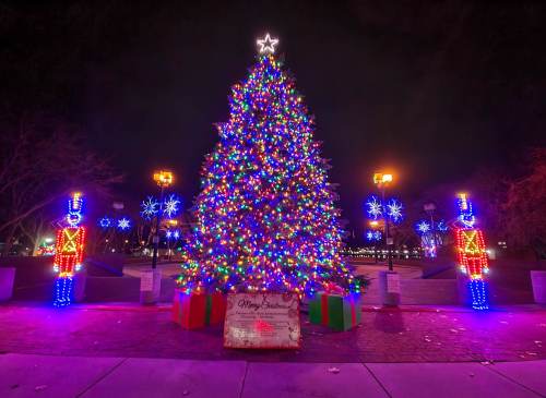 A brightly lit Christmas tree surrounded by colorful lights, gift boxes, and nutcracker figures at night.