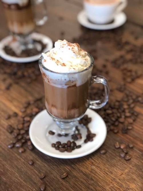 A glass of hot chocolate topped with whipped cream, surrounded by coffee beans on a wooden table.