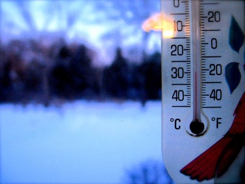 A close-up of a thermometer showing temperatures near freezing, with a snowy landscape blurred in the background.