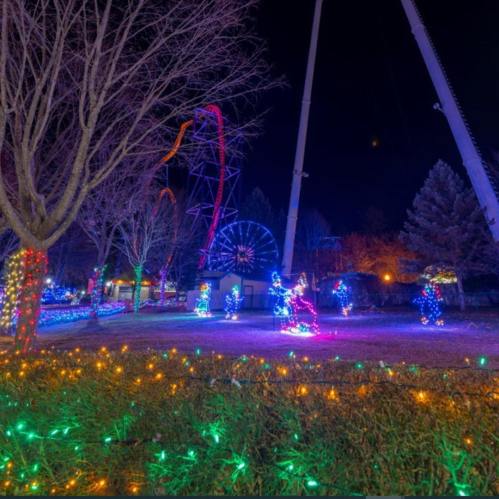 Colorful holiday lights illuminate a park with a ferris wheel and roller coaster in the background at night.