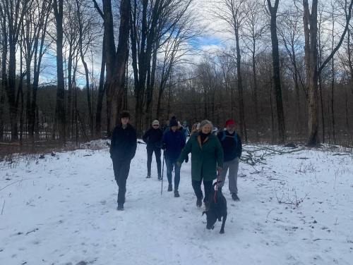 A group of people walking on a snowy path in a forest, accompanied by a dog. Trees are bare against a cloudy sky.