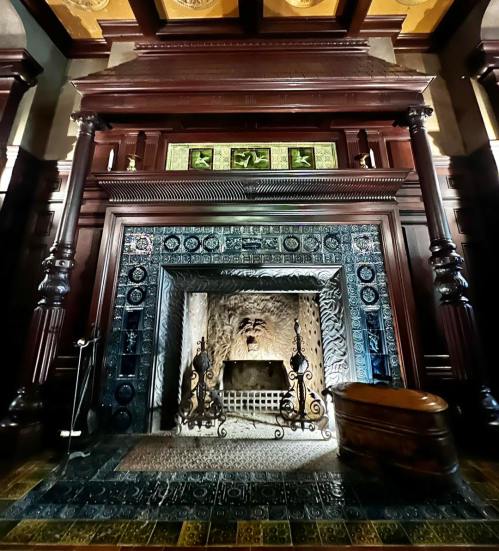 A grand fireplace with intricate tile work and wooden mantel, set in an ornate room with high ceilings.