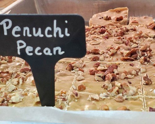 A close-up of a tray of penuchi pecan candy, topped with chopped pecans and displayed with a label.