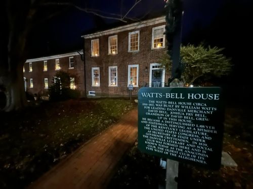 Historic Watts-Bell House at night, featuring brick architecture and illuminated windows, with a sign detailing its history.