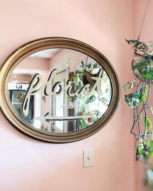 A round mirror with the word "florist" elegantly written on it, surrounded by green plants against a pink wall.