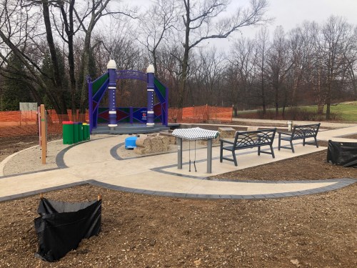 A playground area under construction, featuring benches, a stage, and colorful play equipment surrounded by trees.