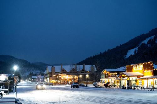 Snow-covered street at dusk, lined with cozy shops and a hotel, surrounded by mountains under a darkening sky.