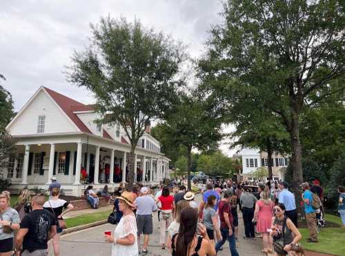 A bustling outdoor event with a crowd gathered around a white house and trees, under a cloudy sky.