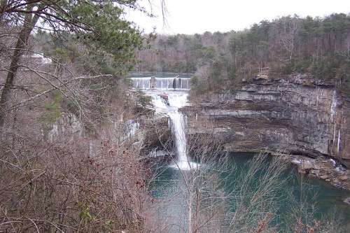 A scenic view of a waterfall cascading into a turquoise pool, surrounded by rocky cliffs and bare trees.