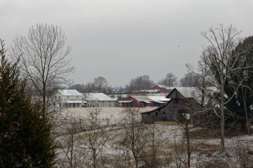 A snowy rural landscape featuring a barn, trees, and distant farm buildings under a gray sky.