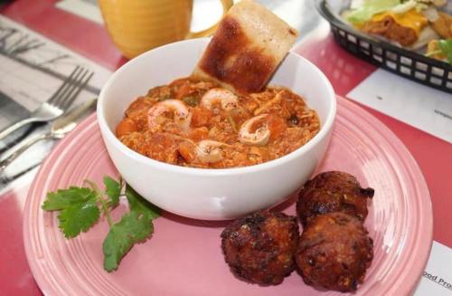 A bowl of shrimp stew with a toasted bread slice, accompanied by two fried balls, on a pink plate.