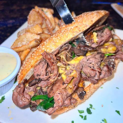 A close-up of a sesame seed sandwich filled with sliced beef, cheese, and greens, served with crispy fries and dipping sauce.