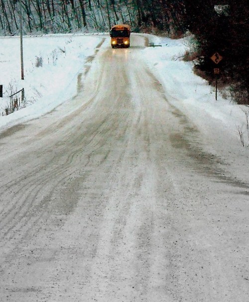 A snow-covered road with a school bus approaching in the distance, surrounded by trees.