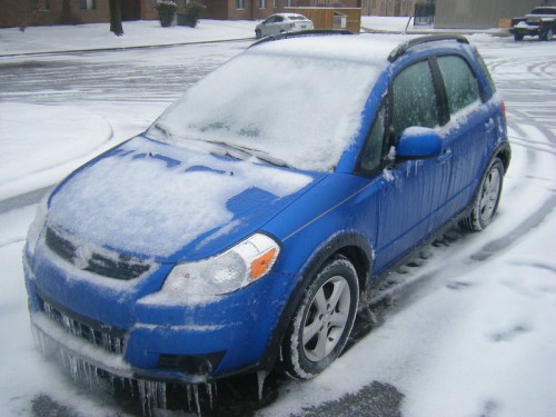 A blue car covered in snow and ice, parked on a snowy street with icicles hanging from it.