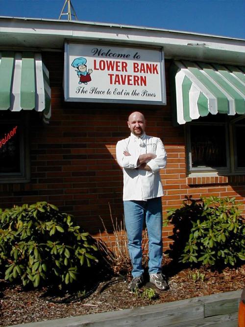 A man in a white chef's coat stands outside the Lower Bank Tavern, with a sign above and greenery around.