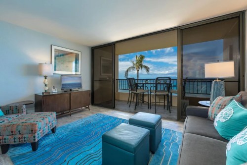 Cozy living room with ocean view, featuring a sofa, chairs, TV, and a balcony with palm trees in the background.
