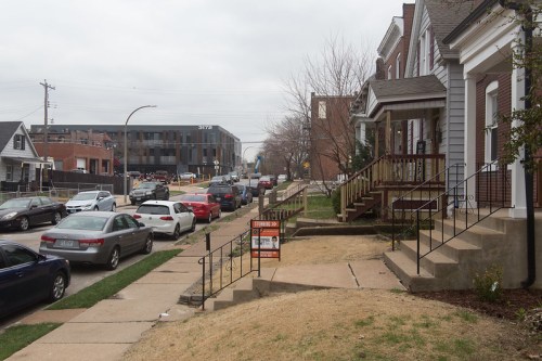 A residential street with parked cars, houses, and a for-sale sign on the lawn, under a cloudy sky.