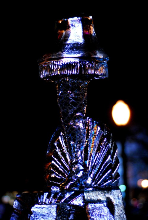 A close-up of a shimmering ice sculpture resembling a lamp, illuminated against a dark background.