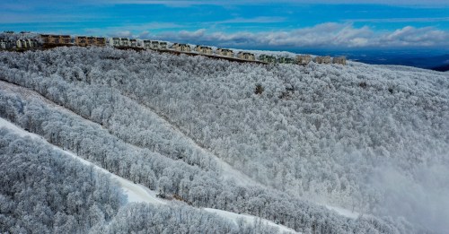 A snowy landscape with frosted trees and a distant cliff under a blue sky, showcasing winter's beauty.