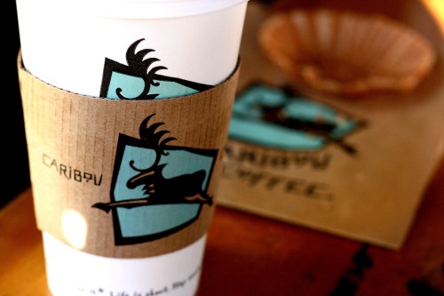 A Caribou Coffee cup with a cardboard sleeve featuring a logo of a caribou, next to a pastry on a plate.