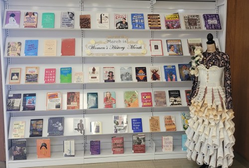 Display celebrating Women's History Month with books on a shelf and a dress made of paper on a mannequin.