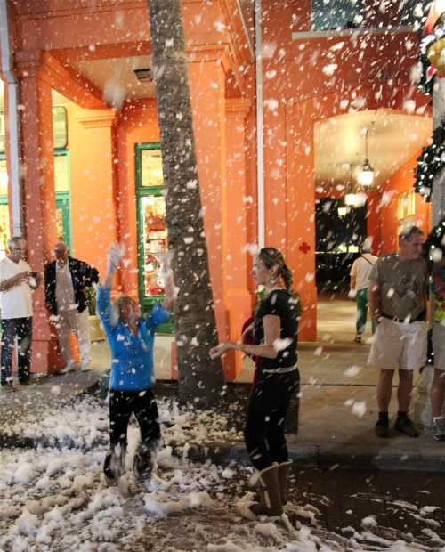 A child joyfully plays in falling snow-like foam while adults watch in a colorful street setting.