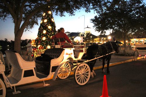 A horse-drawn carriage waits beside a decorated Christmas tree at dusk, with festive lights glowing in the background.