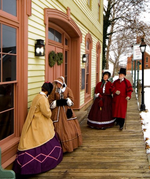 Four people in historical clothing converse outside a colorful building on a snowy day.