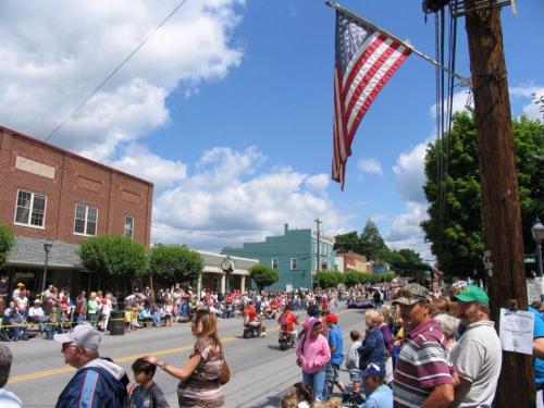 A lively parade scene with a large crowd lining the street, colorful buildings, and an American flag overhead.